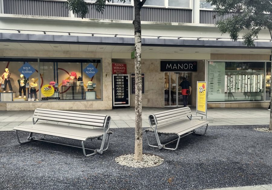Pedestrian shopping street with benches under trees in front of a department store
