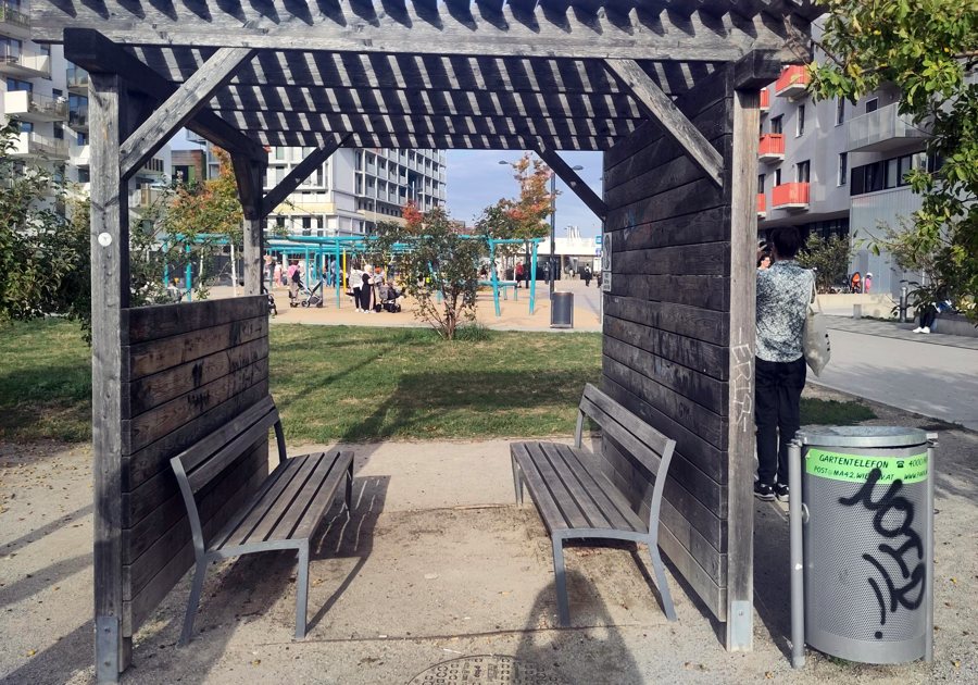 Shaded wooden pergola with benches next to a sunny square